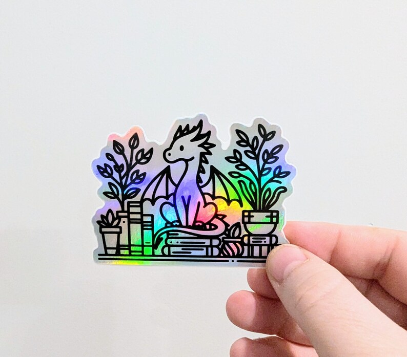 May include: A holographic sticker featuring a dragon sitting on books, surrounded by potted plants. The dragon has a rainbow-colored body and black outlines. The sticker is held by a hand against a white background.