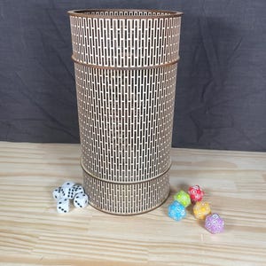 Puede incluir: Una torre de dados cilíndrica alta de madera con un patrón geométrico de ranuras verticales. Varios dados multicolores están dispersos alrededor de la base de la torre, junto con un conjunto de dados blancos con puntos negros. La torre es de madera clara.