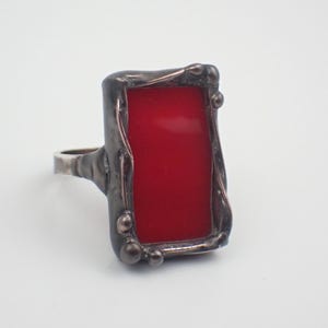 May include: A silver ring with a rectangular, bright red center stone. The stone is framed by a dark, textured metal setting with rounded accents. The ring's band is a simple, polished silver.