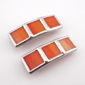 May include: A pair of rectangular hair clips featuring a silver-toned metal frame. Each clip contains three square, vibrant orange inlays with a smooth, polished finish. The design is symmetrical, with the orange segments set side-by-side.