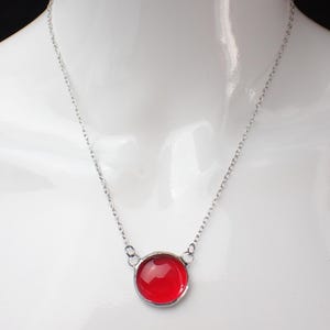 Pode incluir: Colar de corrente prateada com um pendente redondo vermelho brilhante. O pendente é de cor vermelha brilhante e a corrente é delicada. O colar é exibido em um manequim branco.