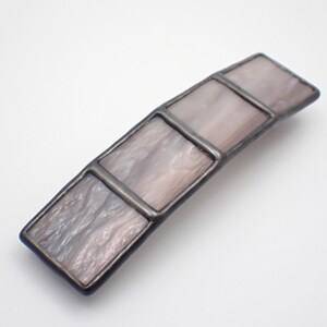 May include: A rectangular hair barrette with a stained glass design. The barrette features four rectangular panels in shades of gray and lavender, framed by a dark metal border. The glass has a marbled appearance.
