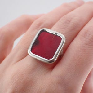 Peut inclure: Une bague dotée d'un élément central carré rouge profond, enchâssé dans une monture en métal argenté lisse et polie. L'insert rouge présente un aspect légèrement texturé et organique avec des variations subtiles de teinte sur sa surface.