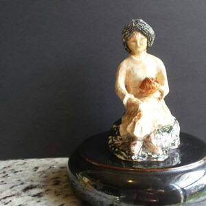 May include: A ceramic figurine of a seated woman holding a basket. The figure is painted in neutral tones with details in brown and black. The figurine sits on a glossy black base, with a black background.