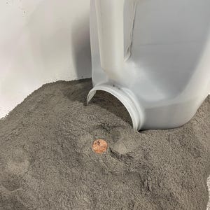 May include: A white plastic jug pouring grey sand into a white container. A penny is visible in the sand.