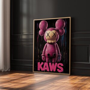 May include: A framed artwork featuring a pink KAWS figure with crossed-out eyes and a skull-like face. The figure has large, rounded ears and gloved hands. The word "KAWS" is printed in pink at the bottom. The artwork is set against a black background.