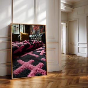 May include: A framed image of a black rug with a pink cross pattern. The rug is folded over, revealing the pattern. The frame is a light brown color. The image is leaning against a white wall in a room with a wooden floor.