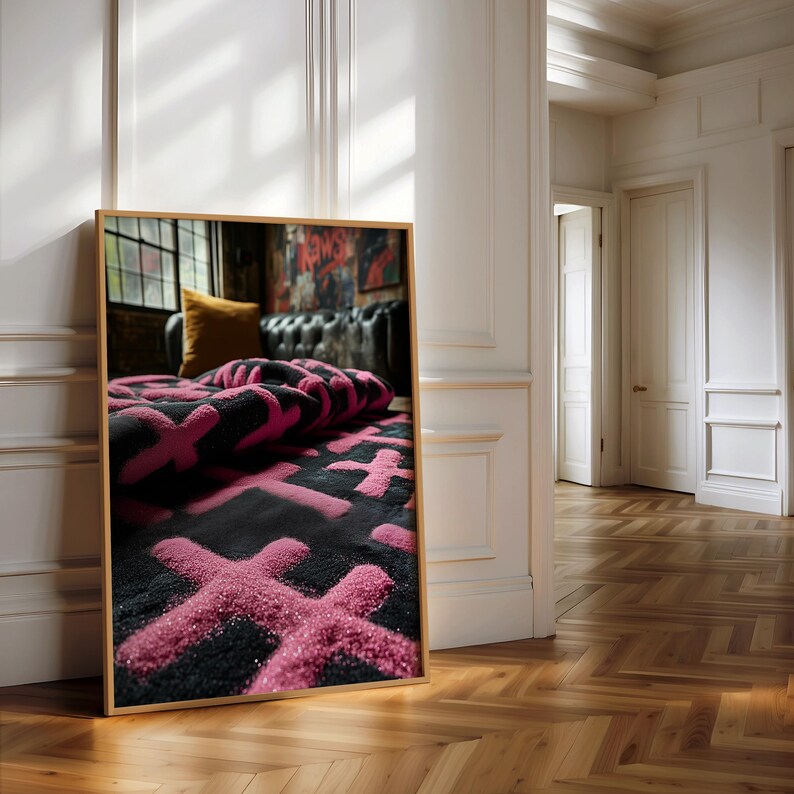 May include: A framed image of a black rug with a pink cross pattern. The rug is folded over, revealing the pattern. The frame is a light brown color. The image is leaning against a white wall in a room with a wooden floor.