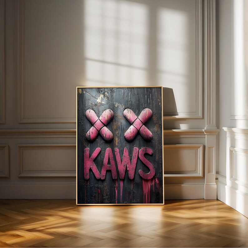 May include: A framed artwork featuring the word "KAWS" in pink, below two pink "XX" shapes. The background is dark with drips of pink paint. The frame is gold, and the artwork is displayed on a wooden floor.