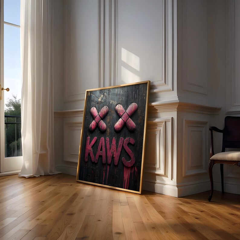 May include: A framed artwork featuring the word "KAWS" in pink, with two pink "XX" symbols above. The artwork has a black background with pink drips and a gold frame. It is leaning against a white wall in a room with hardwood floors.