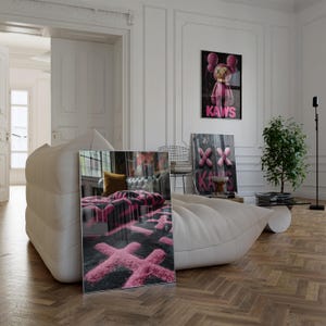 May include: A modern living room with a white leather sofa and a framed artwork featuring a pink and black design. The room has hardwood floors, a small plant, and additional framed art with the word "KAWS".