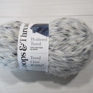 以下が含まれることがあります： ポリエステル混紡のLoops & Threads Heathered Tweedの毛糸玉。白、グレー、黒の斑点模様です。「Loops & Thread」と「Tweed Chiné」のラベルが付いています。