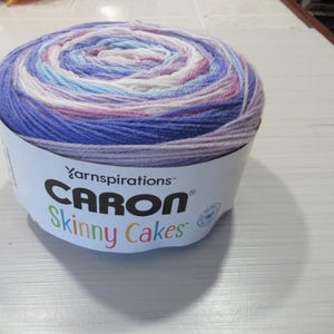 Peut inclure: Pelote de fil Caron Skinny Cakes aux tons de violet, bleu, rose et blanc. Le fil est enroulé en spirale et porte les noms de marque Caron et Skinny Cakes.