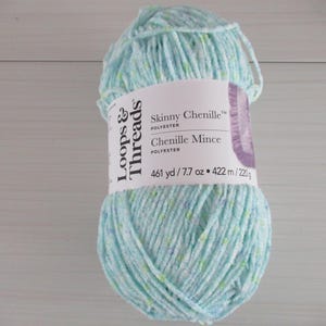Puede incluir: Una madeja de hilo Loops & Threads Skinny Chenille en el color Chenille Mince. El hilo es de color azul claro con motas verdes y blancas. La madeja está etiquetada con el nombre del hilo, el peso y el metraje. El hilo tiene 461 yardas y 7,7 onzas.