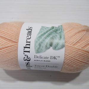 May include: A skein of peach-colored yarn with a white label. The label features the text "Loops & Threads" and "Delicate DK Acrylic Blend". A light blue knitted swatch is also shown on the label. The yarn is for knitting and crochet projects.