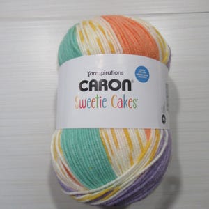 Puede incluir: Ovillo de lana Caron Sweetie Cakes con una mezcla de colores: verde menta, amarillo, naranja y morado. La etiqueta indica "Caron Sweetie Cakes", sugiriendo una lana suave y multicolor, ideal para tejer y hacer ganchillo. Perfecto para proyectos creativos.