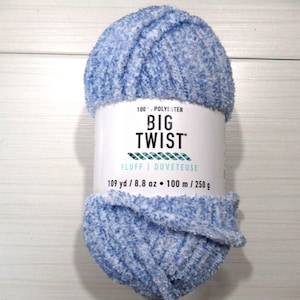 May include: A skein of Big Twist Fluff yarn in a variegated light blue and white colorway. The label indicates 100% polyester, 109 yards, and 8.8 ounces. The yarn has a soft, fuzzy texture and is wrapped in a white paper band with black text.