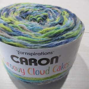 May include: A skein of Caron Groovy Cloud Cakes yarn in a mix of blue, green, and yellow colors. The yarn is in a white paper container with the brand name and product name printed on it.