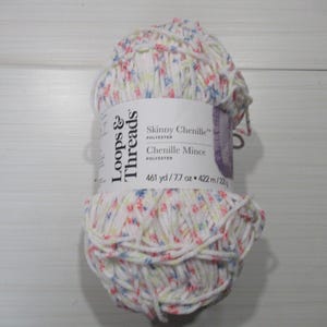 Puede incluir: Una madeja de hilo de chenilla blanco con un estampado floral en rosa, azul y amarillo. La etiqueta dice "Skinny Chenille" y "Chenille Mince" con medidas de 422 metros y 218 gramos.