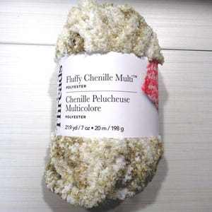 May include: A skein of Fluffy Chenille Multi polyester yarn in a variegated blend of cream, white, and tan. The white paper label displays the product name, material, and measurements of 21.9 yd, 7 oz, 20 m, and 198 g in black text.