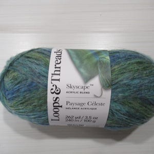May include: A skein of Loops & Threads Skyscape acrylic blend yarn. The yarn is a mix of blue and green hues, with a white label wrapped around it. The label reads "Skyscape" and "Paysage Céleste". The yarn weighs 3.5 oz.