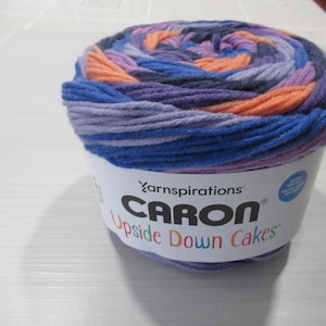 May include: A ball of Caron Upside Down Cakes yarn, featuring a blend of blue, purple, and orange hues. The yarn is wrapped around a white label with the brand and product name in black text.