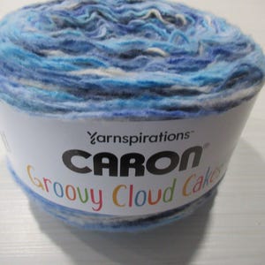 Puede incluir: Ovillo de lana Caron Groovy Cloud Cakes en tonos azules, blancos y grises. La lana está envuelta con una etiqueta que dice "Caron Groovy Cloud Cakes" en texto colorido.