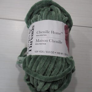 May include: A skein of sage green Loops & Threads Chenille Home polyester yarn. The white product label features the text Chenille Home, Maison Chenille, 109 YDS, 8.8 OZ, 100 M, and 250 G. The yarn has a thick, soft, velvet-like texture.