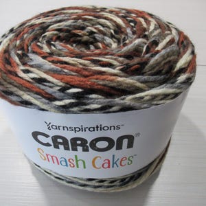 May include: A large ball of Caron Smash Cakes yarn, featuring a mix of brown, orange, grey, and cream colours. The label displays "Yarnspirations CARON Smash Cakes". Suitable for knitting and crochet crafts.