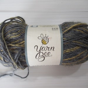 May include: A skein of Yarn Bee yarn in shades of gray and yellow. The label features the brand name and a bee logo. The yarn is tightly wound, ready for knitting or crocheting projects.