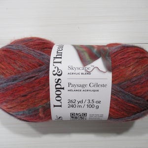 May include: A ball of Loops & Threads Skyscape acrylic blend yarn in shades of red, orange, and gray. The label reads "Paysage Céleste" and indicates 262 yards and 3.5 oz. of yarn. The yarn is on a white surface.