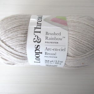 May include: A skein of Loops & Thread "Brushed Rainbow" polyester yarn. The yarn is a light, neutral color with a label that includes the text "Arc-en-ciel Brossé". The label also indicates the yarn is 364 yards / 5.3 oz.