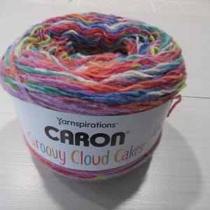 Puede incluir: Una madeja de hilo Caron Groovy Cloud Cakes en una variedad de colores, incluyendo rosa, naranja, azul y amarillo. El hilo está envuelto alrededor de una etiqueta blanca con el nombre de la marca y del producto.