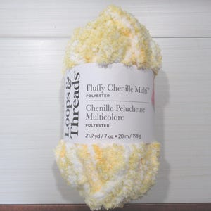 May include: A skein of Loops & Threads Fluffy Chenille Multi polyester yarn in a blend of white and yellow. The white paper label displays the product name, material, and the weight of 7 oz and length of 21.9 yd.