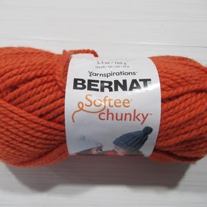 May include: A large ball of orange Bernat Softee Chunky yarn. The label indicates the yarn is 3.5 oz / 100 g and 108 yds / 99 m. The yarn is a solid, vibrant orange color, perfect for knitting or crocheting projects.