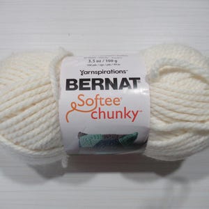 May include: A skein of off-white Bernat Softee Chunky yarn. The label indicates 3.5 oz / 100 g. The yarn is tightly wound, showing a thick, textured appearance. The label features the Yarnspirations logo and a small image of a knitted item.