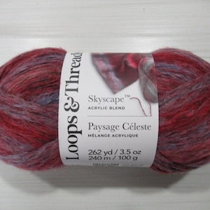 May include: A skein of Loops & Thread Skyscape acrylic blend yarn. The yarn is a mix of red, purple, and gray colors. The label reads "Paysage Céleste" and lists the yarn as 262 yards / 3.5 oz.