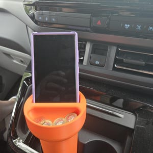 Op de afbeelding: Een oranje bekerhouder voor in de auto met een mobiele telefoon en munten erin. De telefoon zit in een paarse hoes. De bekerhouder zit in de middenconsole van een voertuig.