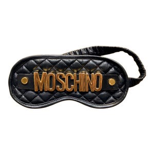 Może przedstawiać: Czarna pikowana maska na oczy ze złotym napisem "MOSCHINO" z przodu. Maska na oczy ma czarny elastyczny pasek. Po obu stronach napisu znajdują się dwa złote nity.