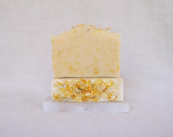 May include: Two bars of handmade soap, one with a pale yellow colour and the other with a white base and yellow flower petals. The soap bars are on a white marble surface.