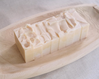 Puede incluir: Una pila de barras rectangulares de jabón blanco en una bandeja de madera. El jabón está dispuesto en forma de espiga, con las barras superponiéndose ligeramente.