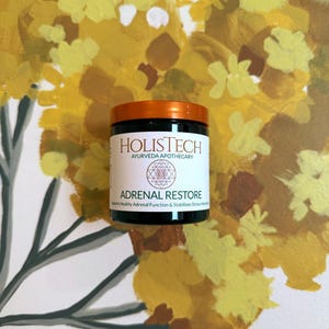 May include: A black jar with an orange lid, labeled "HOLISTECH AYURVEDA APOTHECARY" and "ADRENAL RESTORE." The jar is set against a backdrop of a yellow and brown floral painting. The text on the label states that it supports healthy adrenal function and stabilizes stress hormones.