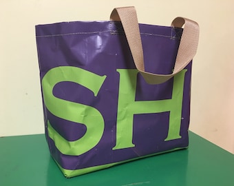 May include: A purple tote bag with the letters "SH" in bright green. The bag has beige handles and is made of a durable, possibly waterproof material. The bag is designed to be reusable and is suitable for carrying groceries or other items.