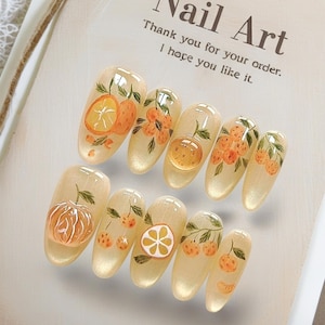 May include: A set of artificial nails with a light yellow base, featuring hand-painted orange fruit designs. The nail art includes oranges, orange slices, and orange blossoms. The text on the tag reads "Nail Art" and "Thank you for your order. I hope you like it."