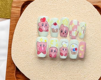May include: A set of ten press-on nails with a light green base, each decorated with cartoon character designs in pink, red, and white. Designs include hearts, cupcakes, stars, and a checkered pattern. The nails are arranged on a beige surface.