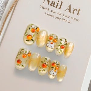 Puede incluir: Juego de uñas artificiales con base transparente, con diseños pintados a mano de naranjas en ramas y un gato blanco con corona. Las uñas tienen un acabado brillante y están dispuestas sobre una tarjeta blanca con el texto "Nail Art".