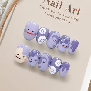 May include: A set of lavender-colored artificial nails with cartoon character designs. The nails feature various designs, including smiling faces, and graphic patterns. The card behind the nails reads "Nail Art" and "Thank you for your order. I hope you like it."