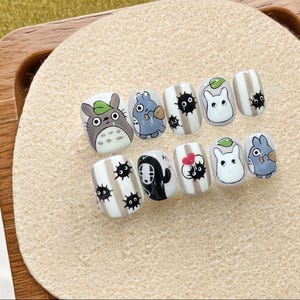 Puede incluir: Un conjunto de uñas postizas con diseños de personajes de dibujos animados. Las uñas presentan un Totoro gris, criaturas azules y grises, conejos blancos con hojas verdes y duendes de hollín negros. Algunas uñas tienen rayas beige verticales.