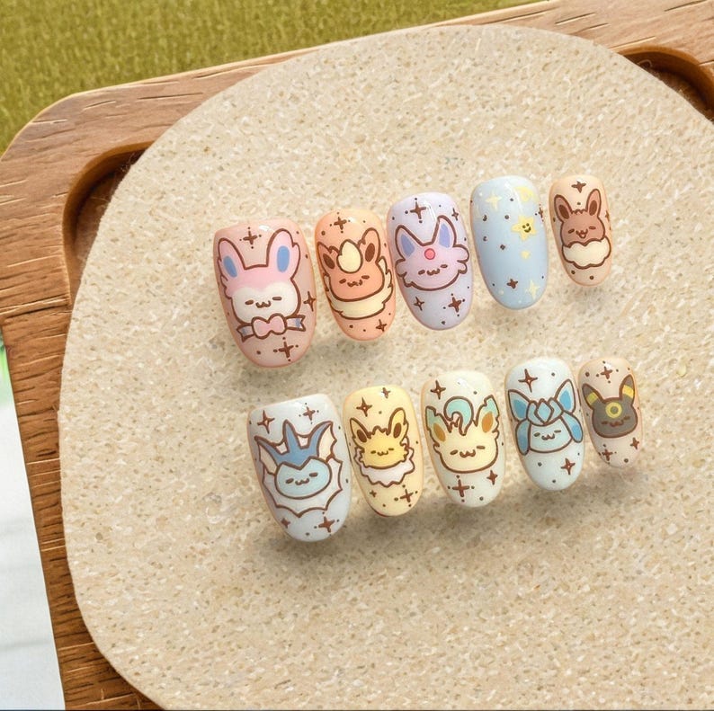 May include: A set of ten press-on nails featuring various pastel-colored designs with cartoon character faces, small stars, and sparkles. The nails are arranged in two rows, showcasing shades of pink, peach, lavender, light blue, yellow, and cream.
