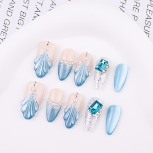 Könnte beinhalten: Eine Sammlung künstlicher Fingernägel in verschiedenen Designs. Einige Nägel zeigen ein hellblaues Muscheldesign, andere einen großen, quadratischen, blauen Edelstein, und einige sind einfarbig hellblau. Die Nägel sind auf weißem Hintergrund.
